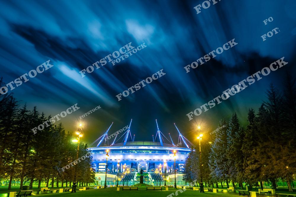 На фото — стадион «Газпром Арена» (также известный как «Санкт-Петербург Арена»), одно из крупнейших спортивных сооружений России. Здание эффектно подсвечено синим светом, а над ним раскинулось динамичное облачное небо, создающее драматичную атмосферу. На переднем плане — аллея с фонарями и деревьями, ведущая к арене. Стадион расположен на Крестовском острове и вмещает более 60 тысяч зрителей. Здесь проходят футбольные матчи, концерты и масштабные мероприятия.